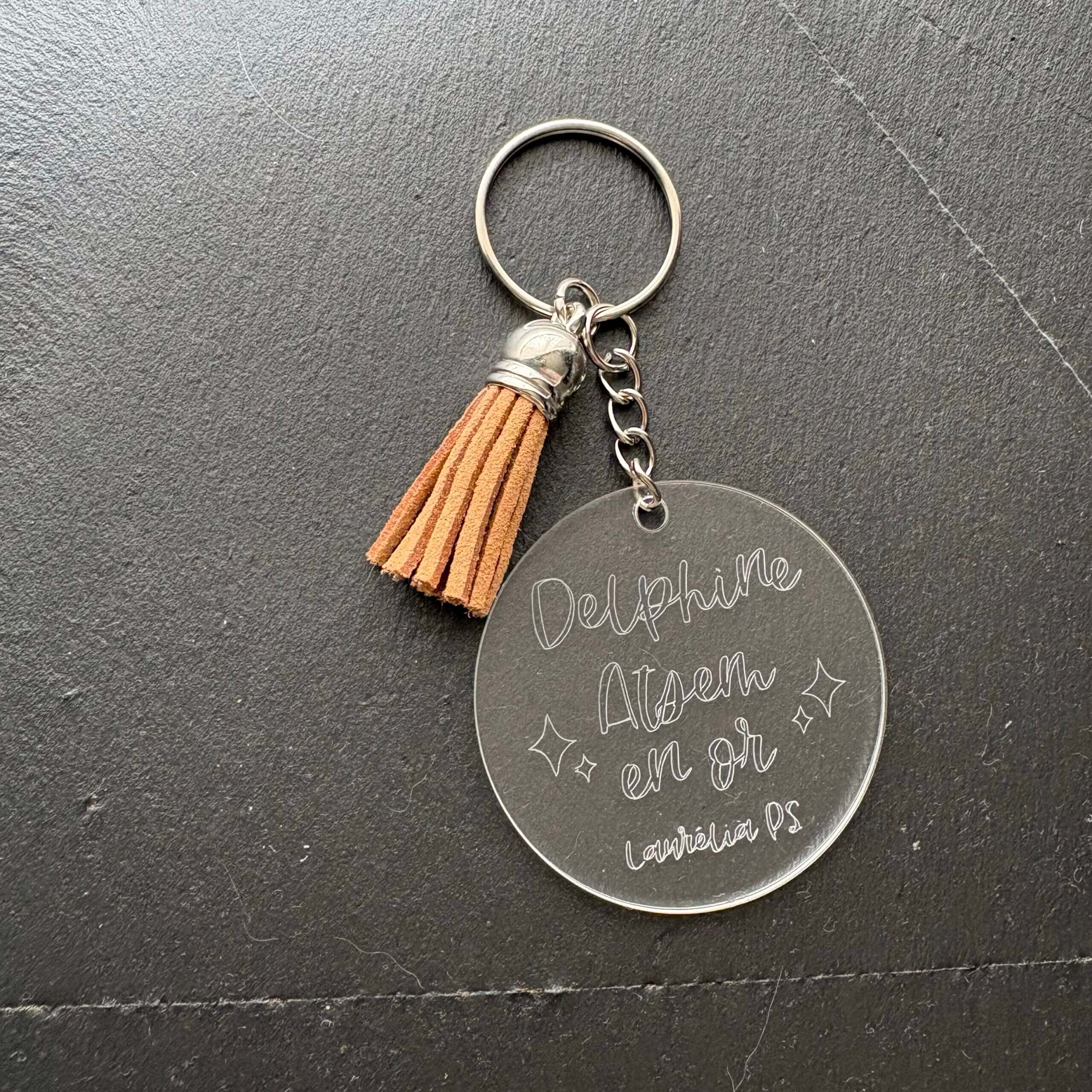 Cadeau personnalisé ATSEM : porte-clés rond transparent gravé Delphine Atsem en or avec pompon en suédine marron.