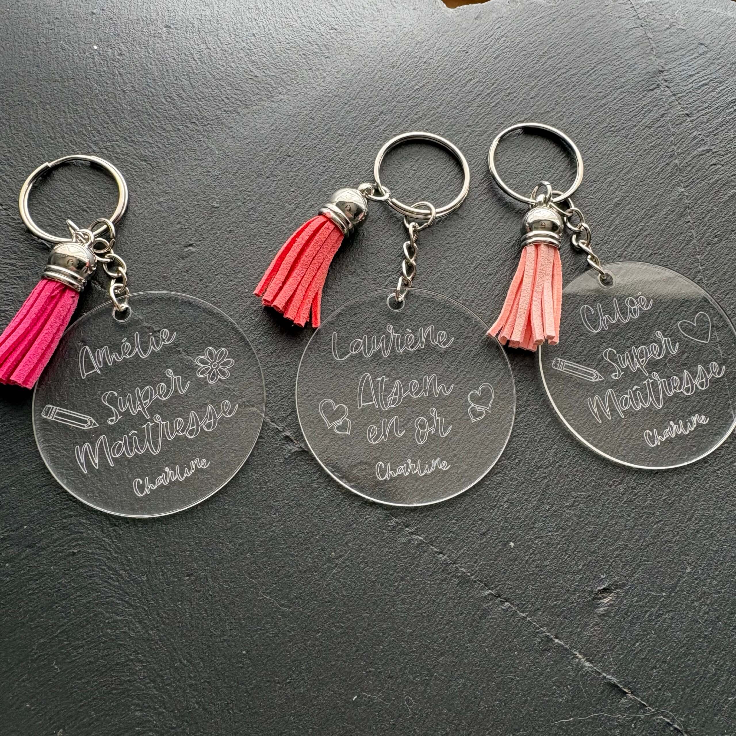 Idée cadeau maîtresse et ATSEM personnalisé. Trois porte-clés transparents gravés avec les prénoms Amélie, Laurène et Chloé et des pompons roses.