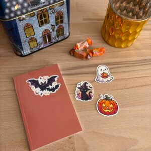 Vue de dessus d'une table en bois décorée pour Halloween. Un carnet terracotta avec un sticker de chauve-souris est posé à côté de stickers d'un chat sorcier, d'un fantôme tenant une citrouille et d'une Jack-o'-lantern. Au second plan, une boîte en forme de maison hantée, un photophore doré et quelques bonbons à la citrouille.