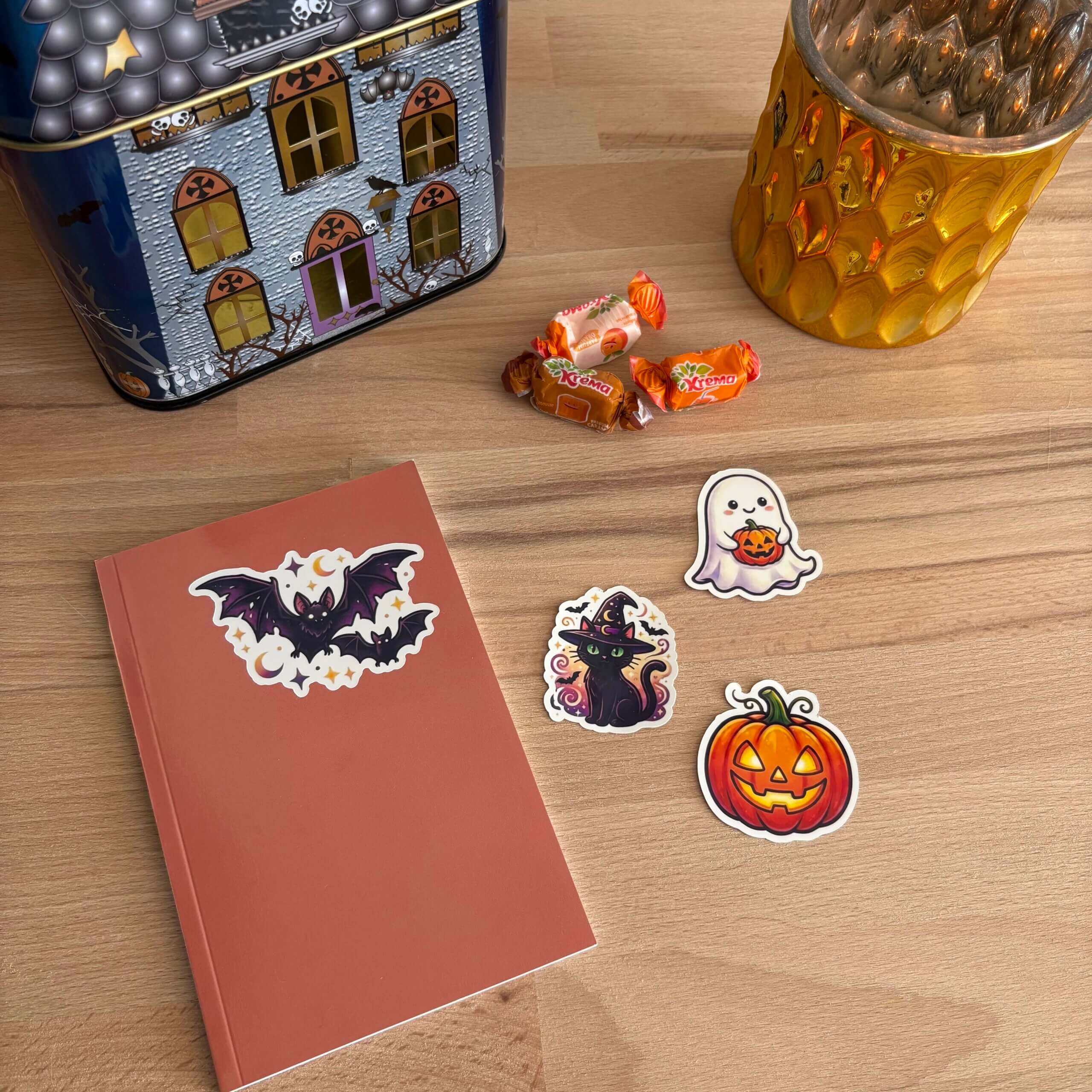 Vue de dessus d'une table en bois décorée pour Halloween. Un carnet terracotta avec un sticker de chauve-souris est posé à côté de stickers d'un chat sorcier, d'un fantôme tenant une citrouille et d'une Jack-o'-lantern. Au second plan, une boîte en forme de maison hantée, un photophore doré et quelques bonbons à la citrouille.