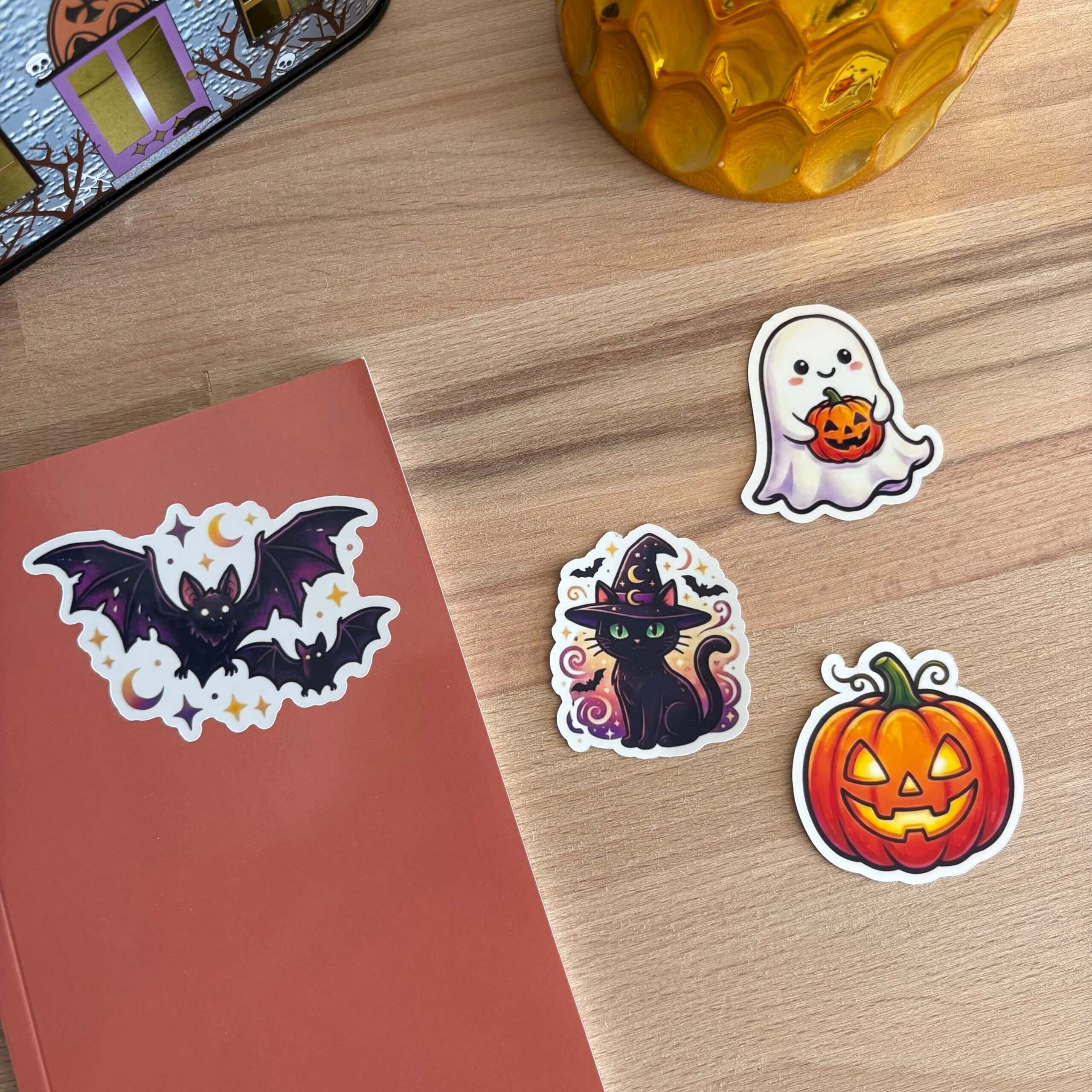 Plan rapproché sur une table en bois montrant le coin d'un carnet de notes terracotta. Un sticker de chauve-souris stylisée est collé sur la couverture du carnet. À côté, les stickers d'un chat noir en chapeau de sorcière, d'un fantôme mignon tenant une mini-citrouille et d'une citrouille d'Halloween souriante sont disposés sur la table.