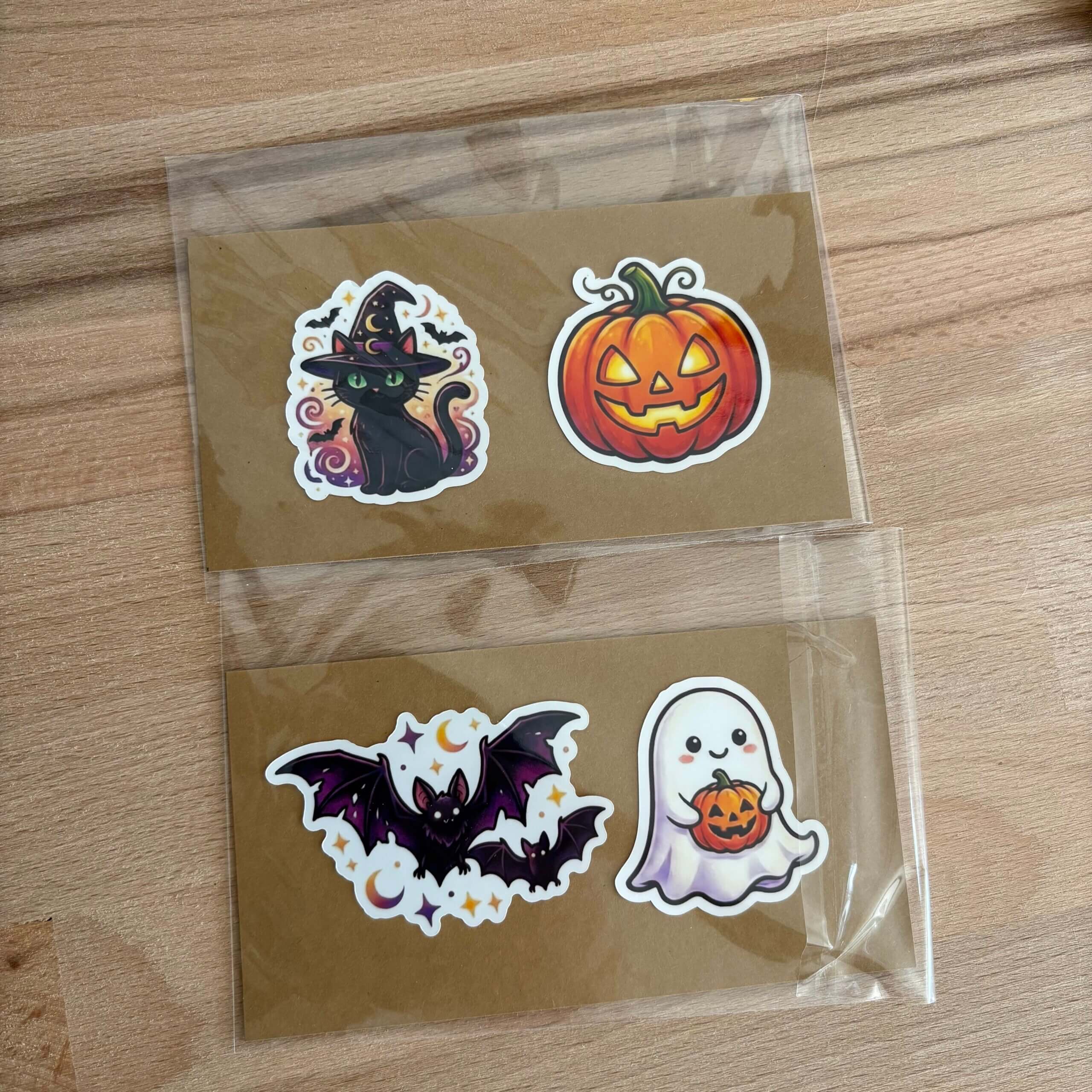 Deux pochettes transparentes contenant des stickers d'Halloween sont posées sur une table en bois. Chaque pochette contient un sticker sur un support en carton craft. La pochette du haut montre le sticker du chat sorcier et de la citrouille. La pochette du bas montre le sticker de la chauve-souris et du fantôme. Les paquets sont scellés et prêts à être expédiés.