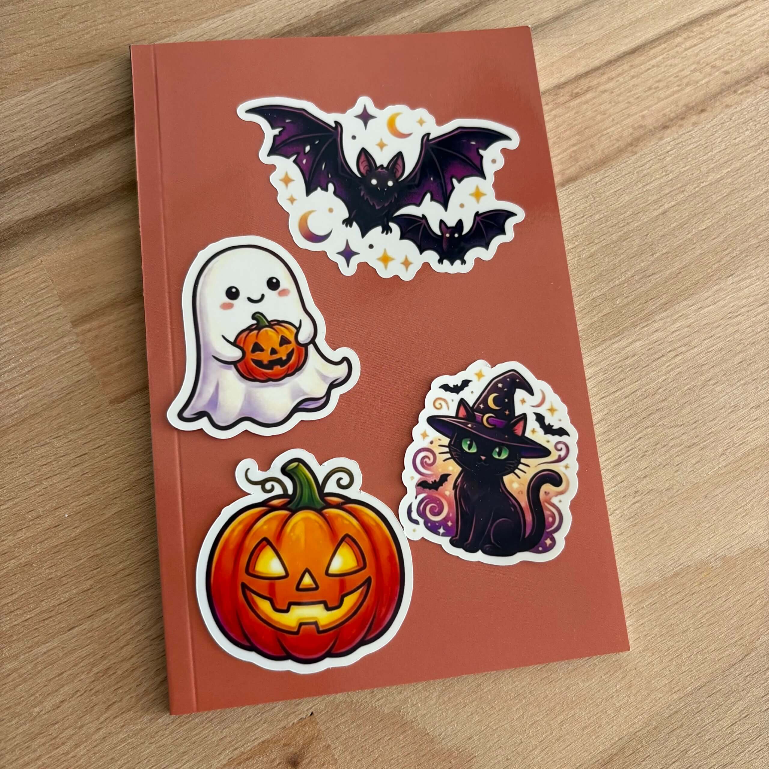 Vue d'ensemble d'un carnet de notes terracotta posé sur une surface en bois. La couverture est décorée avec les quatre stickers de la collection : un grand sticker de chauve-souris en haut, flanqué d'un fantôme tenant une citrouille et d'un chat sorcier au centre, et une citrouille Jack-o'-lantern en bas.