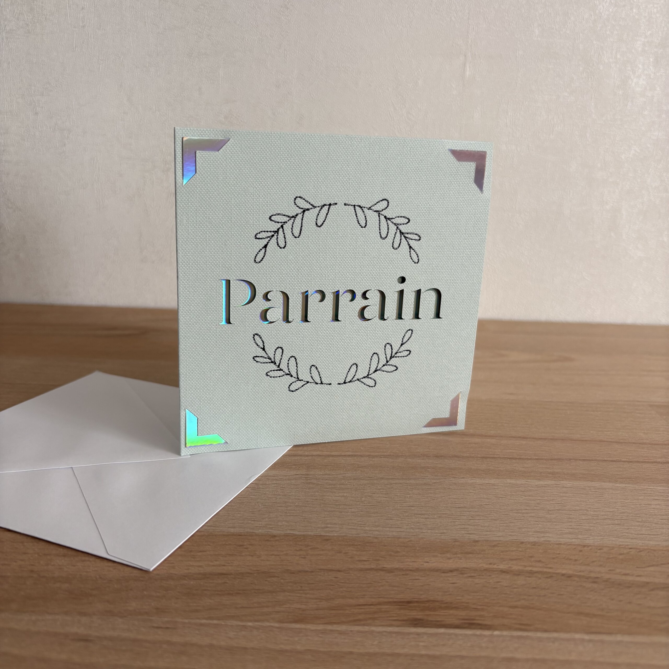 Carte Parrain faite main en papier texturé avec couronne de feuillage et détails holographiques, accompagnée de son enveloppe