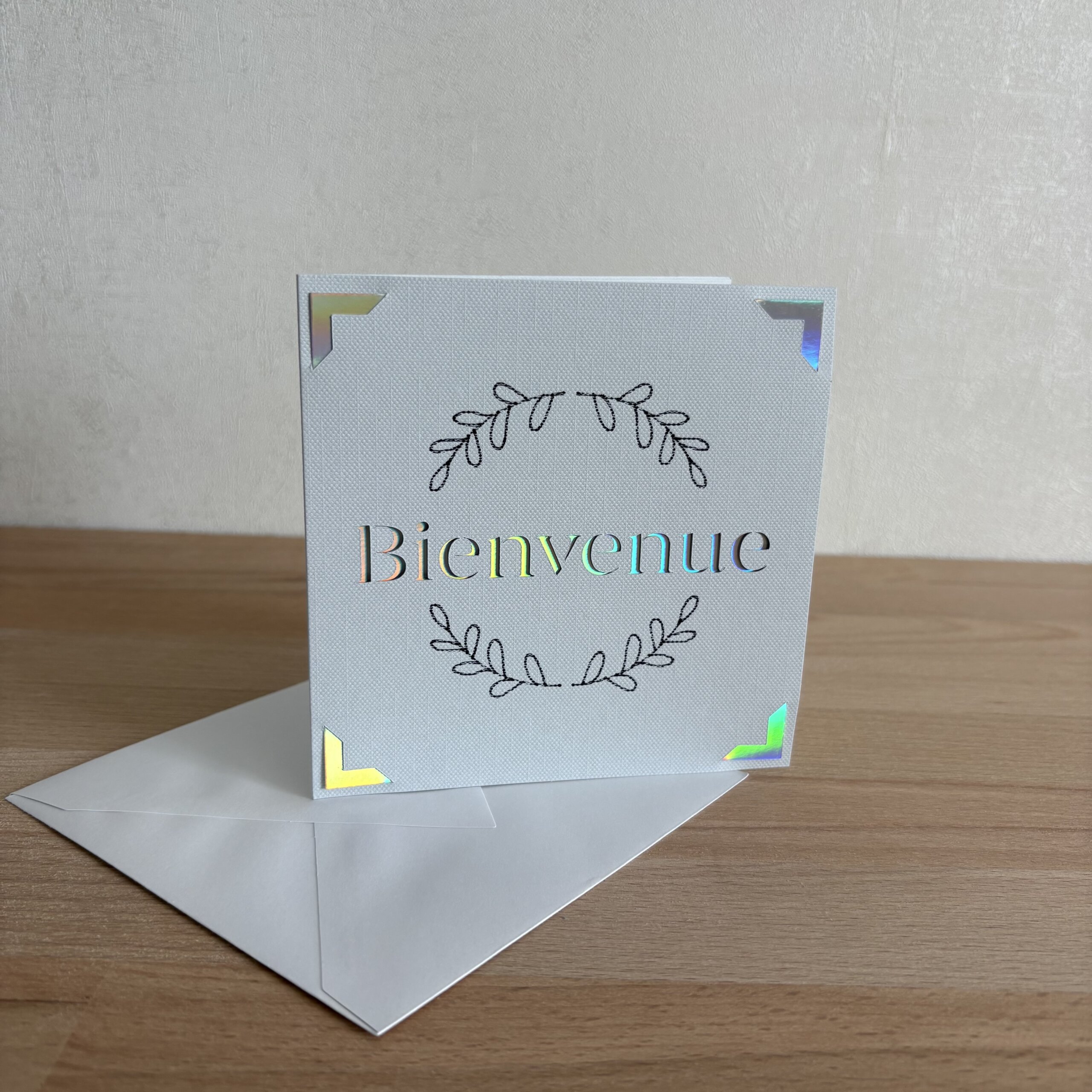 Carte Bienvenue faite main en papier texturé clair avec couronne de feuillage et détails holographiques, accompagnée de son enveloppe
