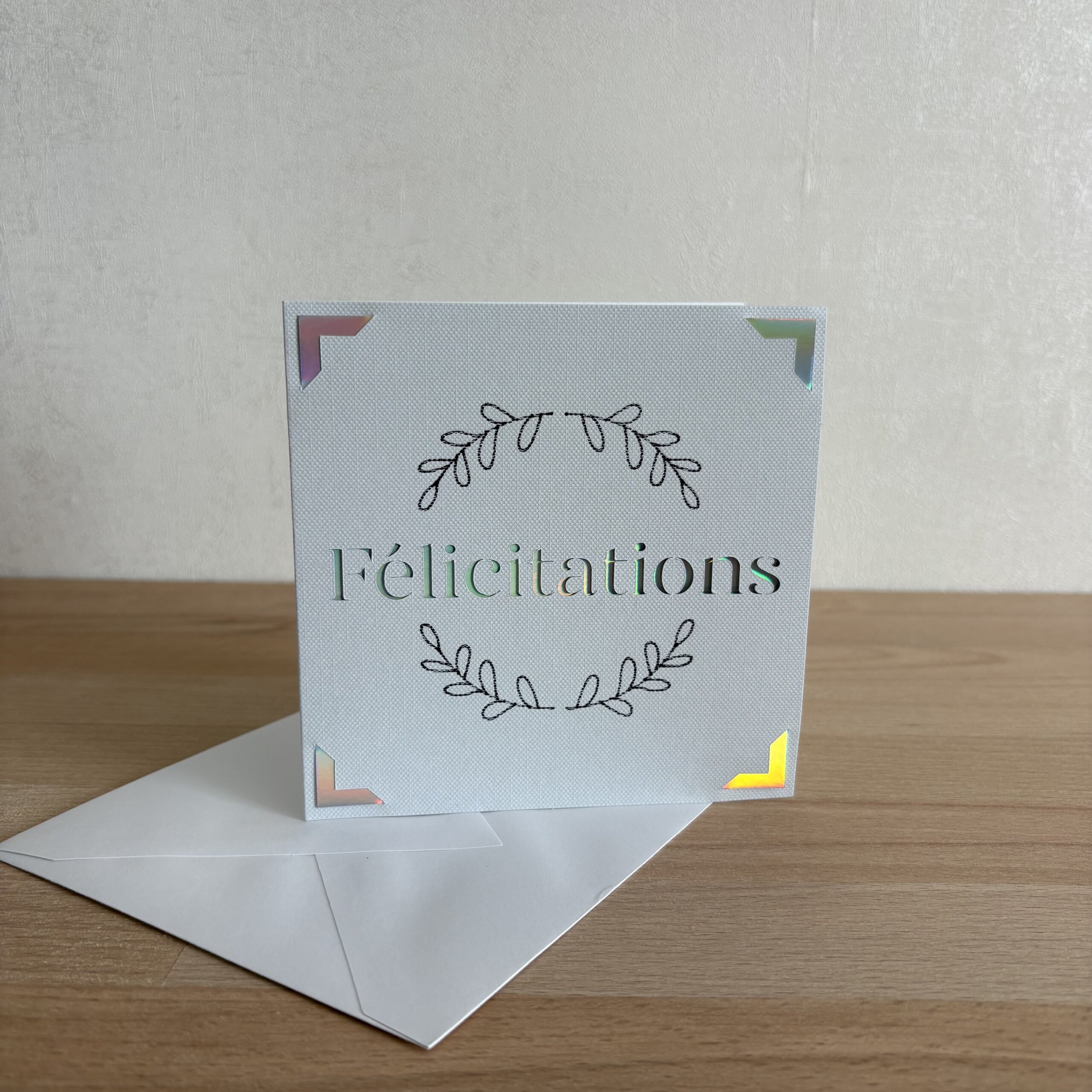 Carte Félicitations faite main en papier texturé clair avec couronne de feuillage et détails holographiques, accompagnée de son enveloppe