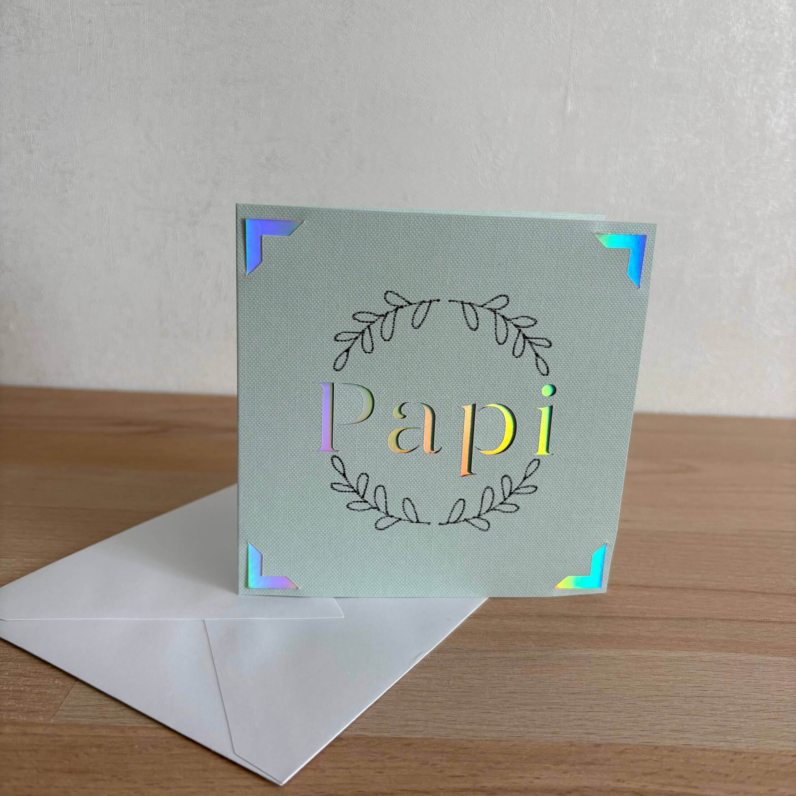 Carte Papi faite main en papier texturé vert clair avec couronne de feuillage et détails holographiques, accompagnée de son enveloppe
