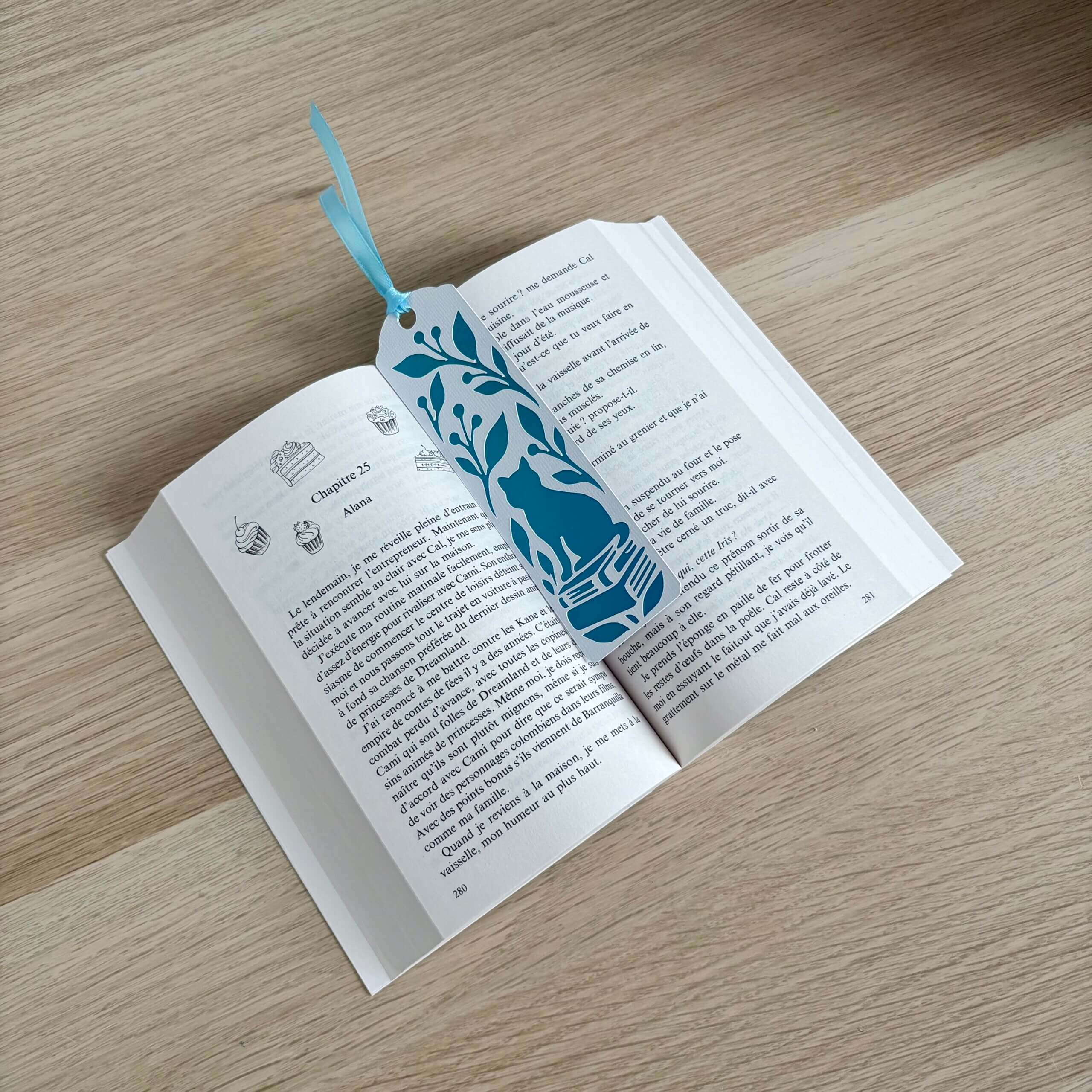 Marque-page chat sur des livres avec feuillage, papier blanc texturé et papier miroir bleu turquoise, présenté dans un livre