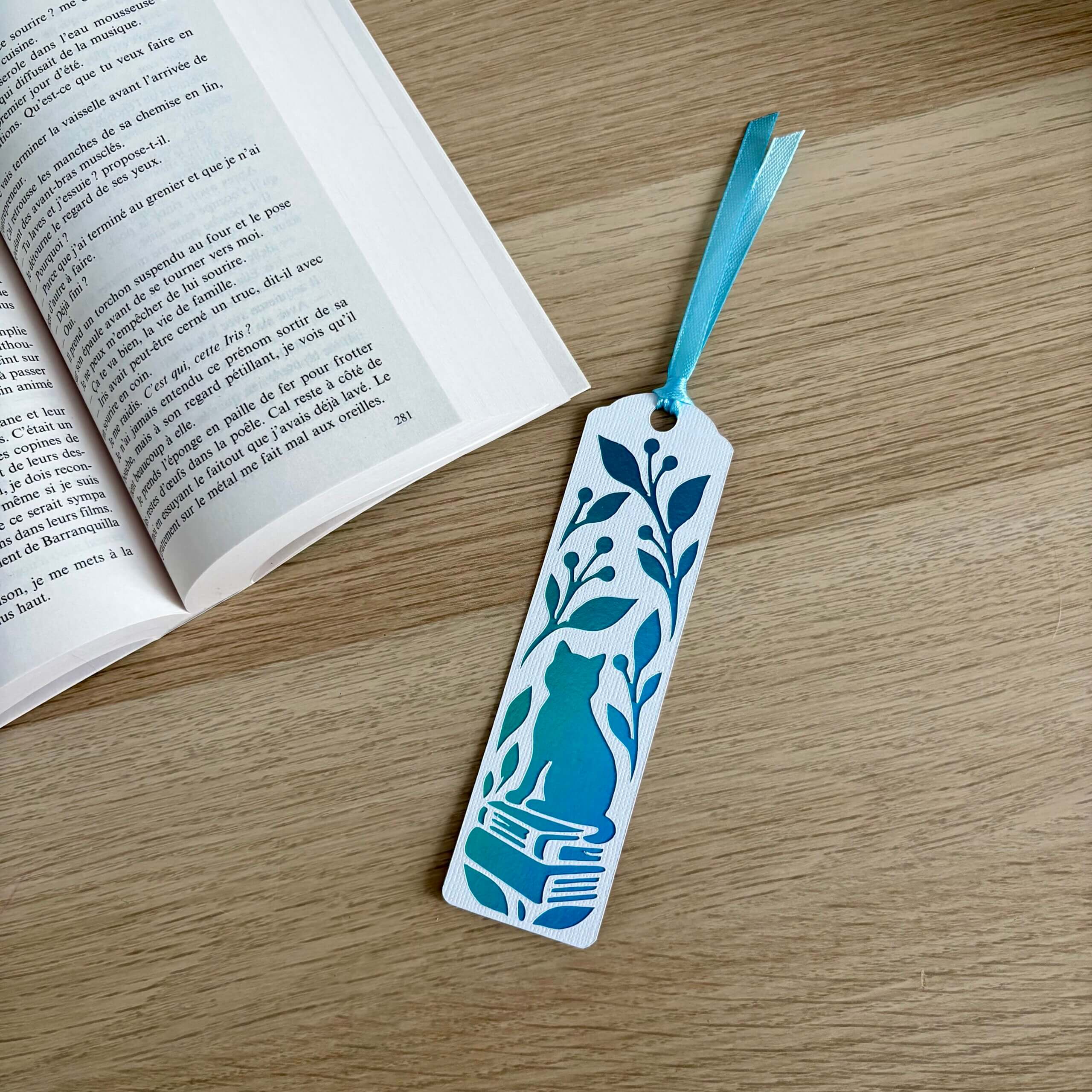 Marque-page ajouré avec chat assis sur des livres et feuillage, en papier blanc texturé et miroir bleu turquoise